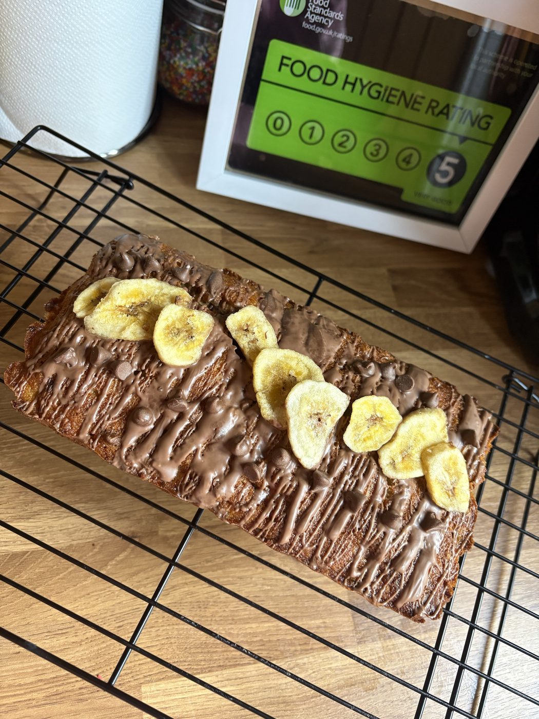 Banana and chocolate chip loaf with a milk chocolate drizzle and banana chips, beside a 5 Food Hygiene Rating certificate