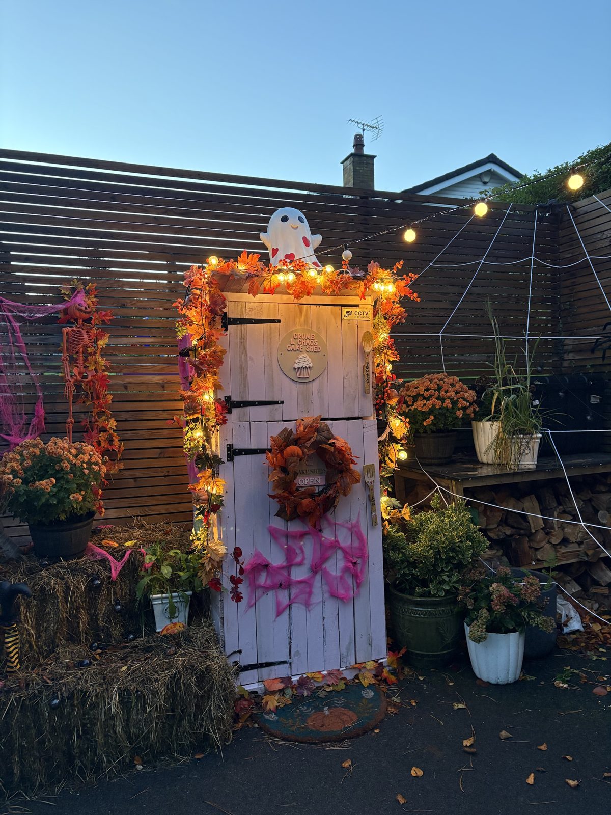 The Crumb & Chaos Cake Shed lit up for Halloween with autumn garland, fairy lights, a ghost on top, hay bales and a giant cobweb
